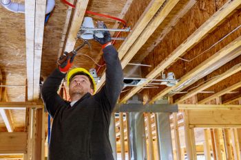 Construction worker installing electrical wiring in an unfinished house interior with exposed ceiling joists